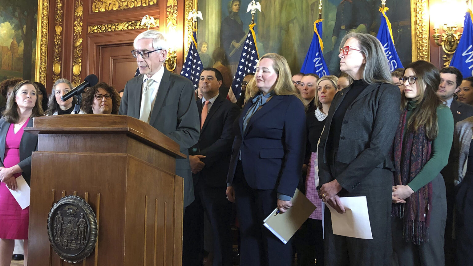 Tony Evers stands behind a podium with a brass Great Seal of the State of Wisconsin and speaks, with Josh Kaul behind him and Sara Rodriguez to his right, standing among state legislators with paintings, gilded wall moulding, and U.S. and Wisconsin flags in the background.