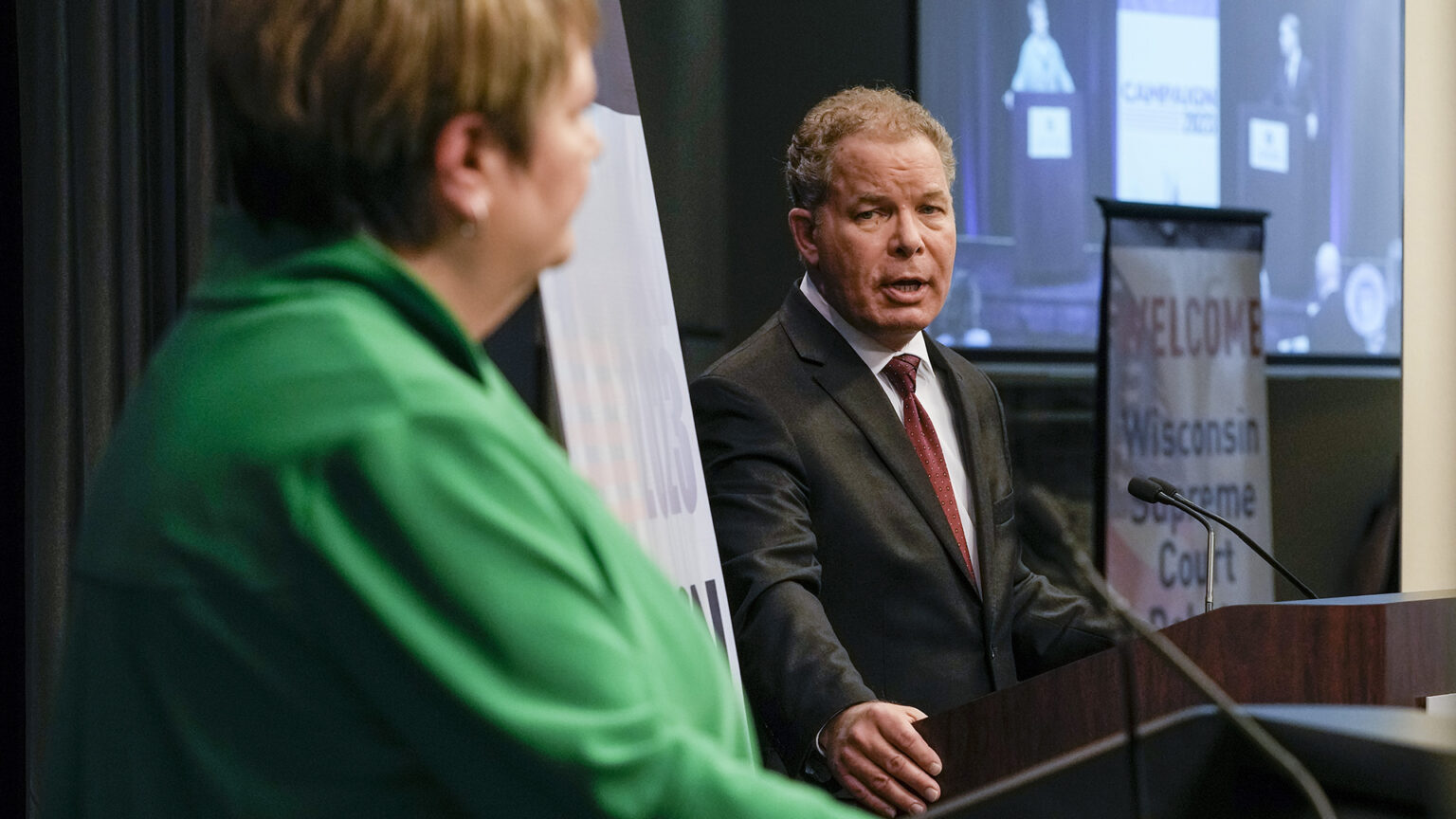 Dan Kelly stands at a podium and speaks to Janet Protasiewicz, standing at another podium in the foreground (and out of focus), with vertical banners and a projection screen displaying a direct angle view of both individuals.