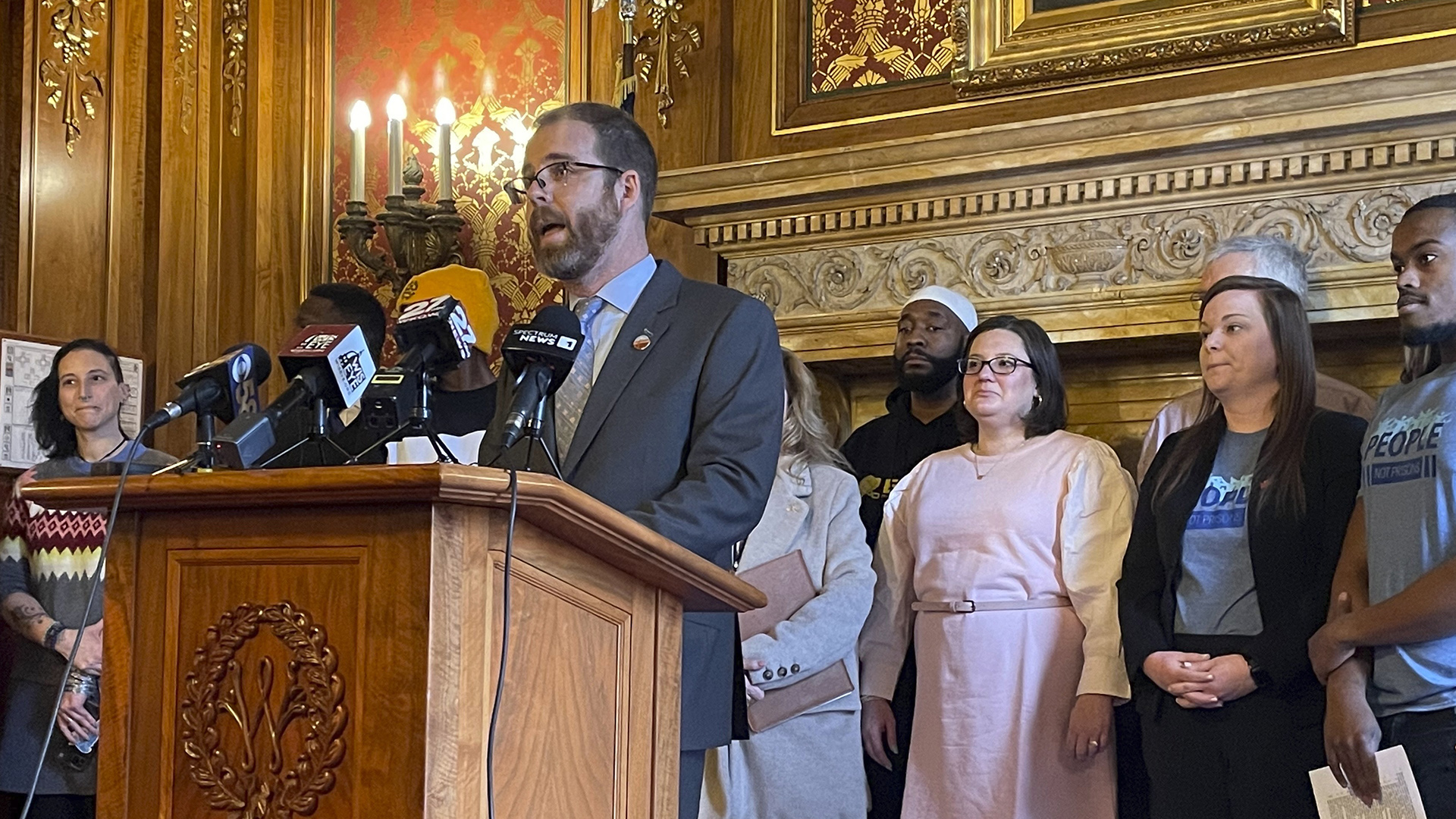 Ryan Clancy speaks while standing behind a podium mounted with multiple microphones, with other people standing behind him in a room with wood moulding, toile wallpaper and electric sconces.