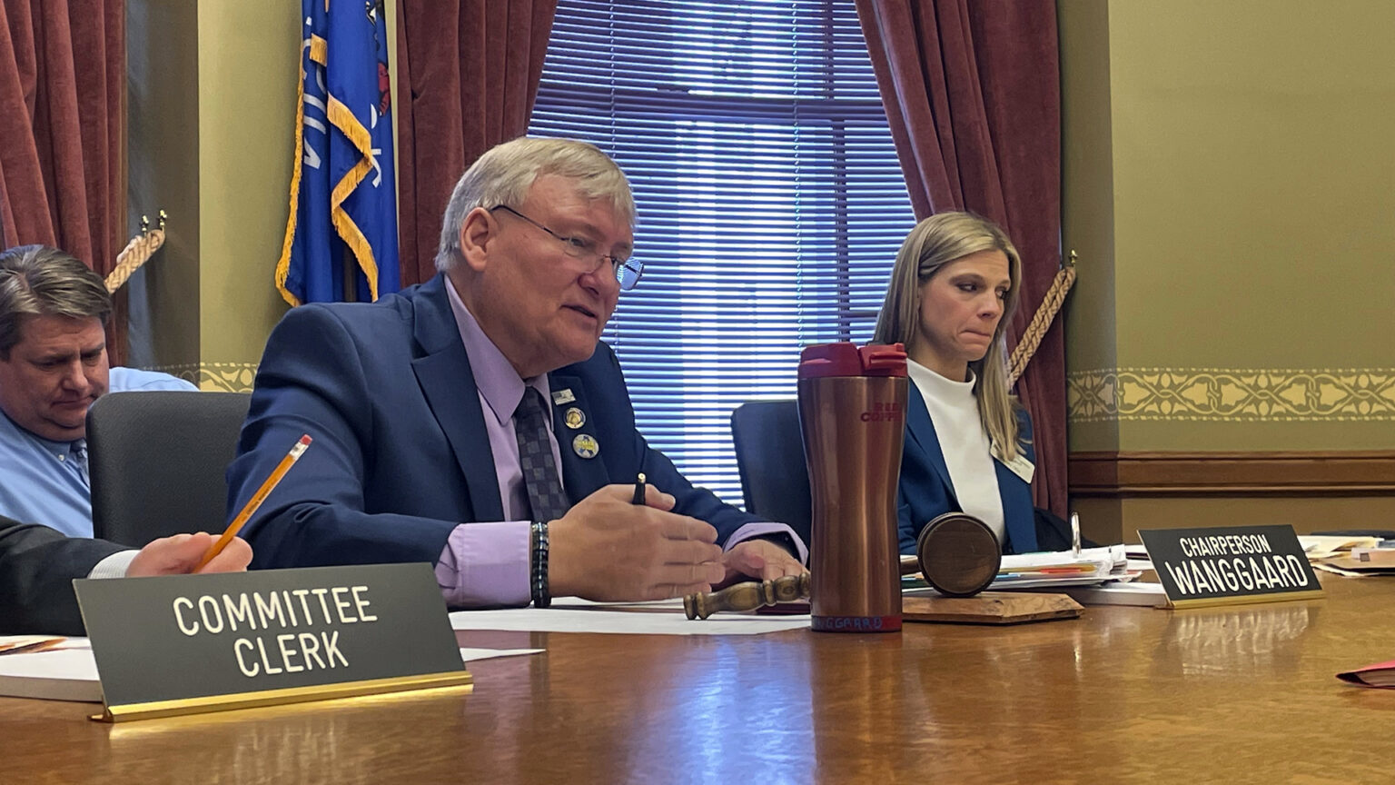 Van Wanggaard speaks and gestures with his right hand while sitting at a table with a nameplate reading Chairperson Wanggaard, an insulated beverage mug and other items, with other people seated to his side and behind him, in a room with a Wisconsin flag and a window with drawn blinds and open drapes.