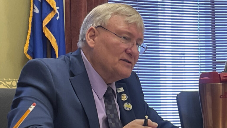 Van Wanggaard speaks while sitting in a room with a Wisconsin flag and a window with drawn blinds and open drapes.