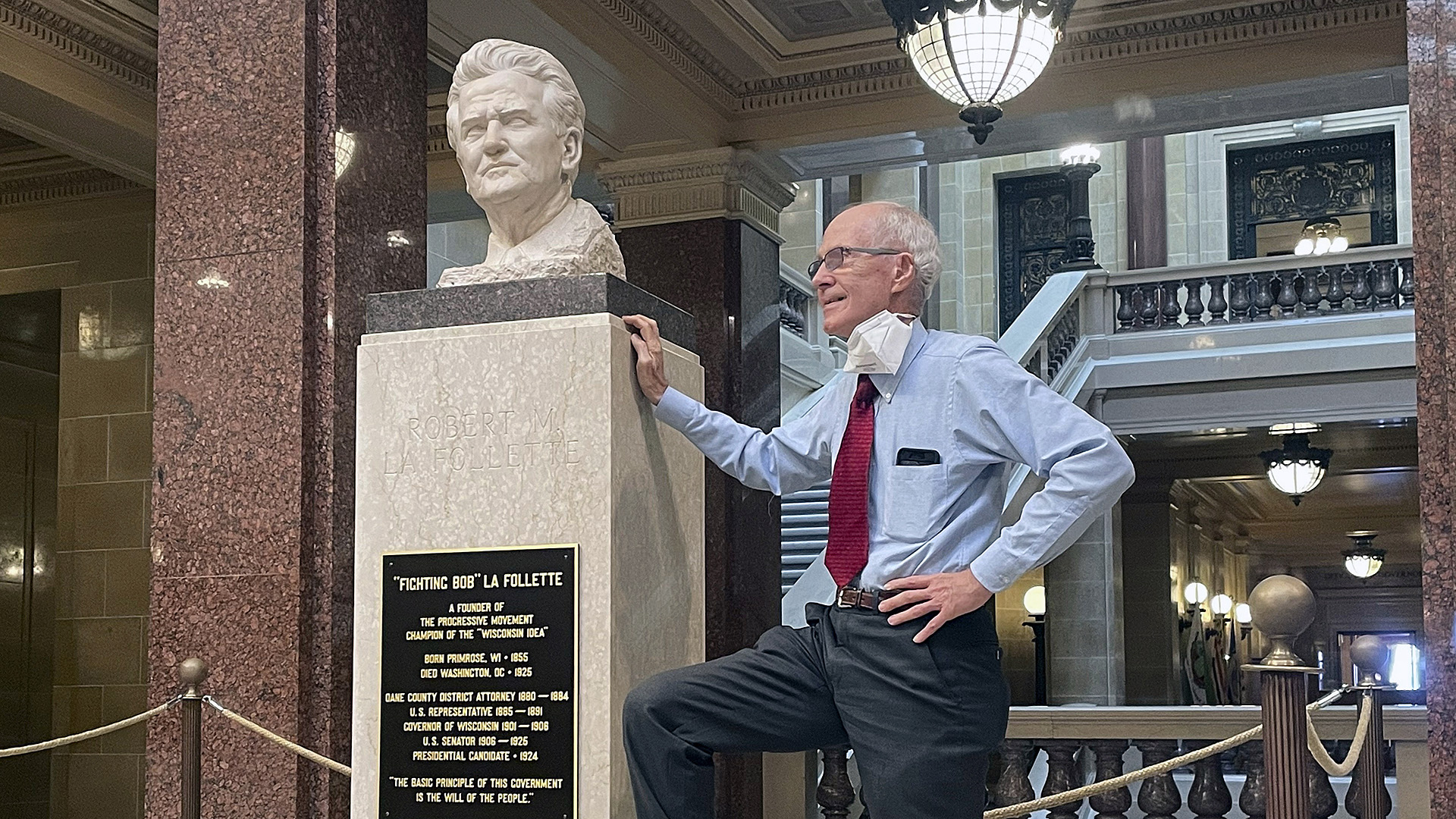 Doug La Follette poses next to a bust of "Fighting Bob La Follette" placed atop a plinth in the rotunda of the Wisconsin State Capitol, with hallways, staircases and bannisters made with marble masonry in the background.