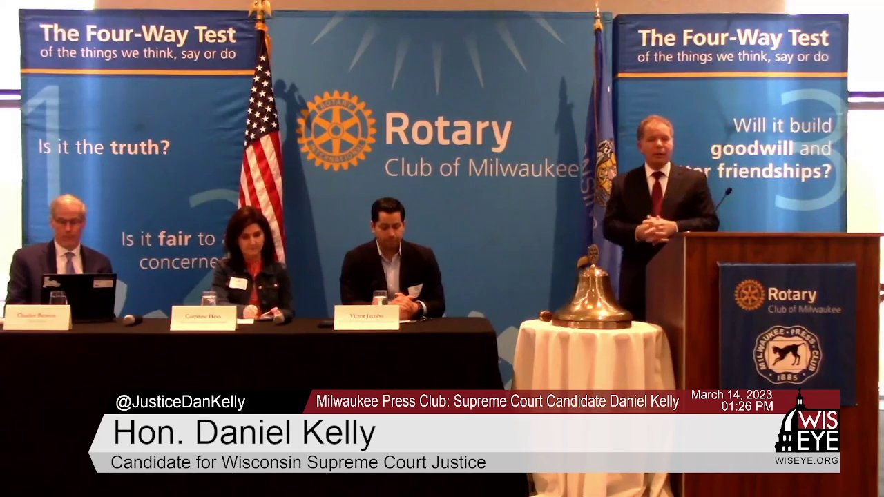 Daniel Kelly stands behind a podium and speaks while three people seated at a table to his right listen and take notes, with the U.S. and Wisconsin flags in the background along with a backdrop banner for the Rotary Club of Milwaukee.