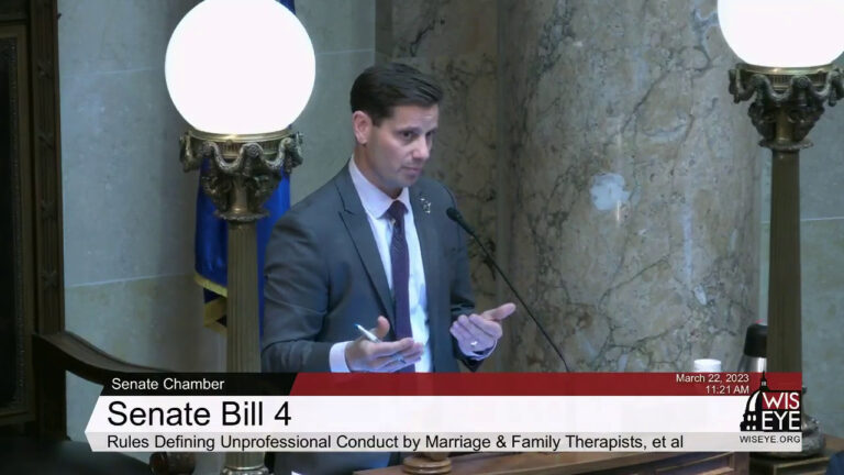 Chris Kapenga stands before a podium with a microphone and gestures with both hands, with two brass light fixtures on either side of him, and with a Wisconsin flag and marble masonry in the background.