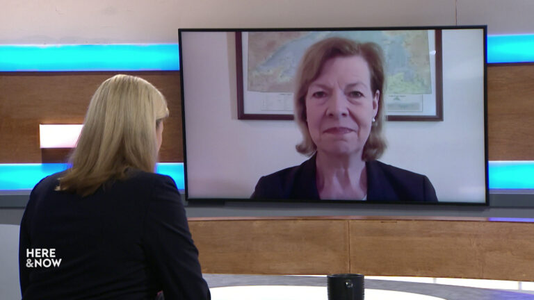 Frederica Freyberg sits at a desk on the Here & Now set and faces a video monitor showing an image of Tammy Baldwin.
