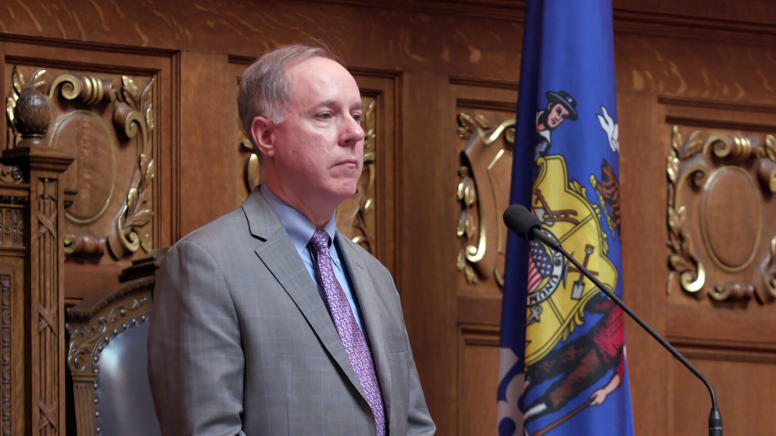 Robin Vos stands from the Speaker's chairs on the dais at the front of the Wisconsin Assembly chamber, with a Wisconsin flag and wood-paneled walls in the background.