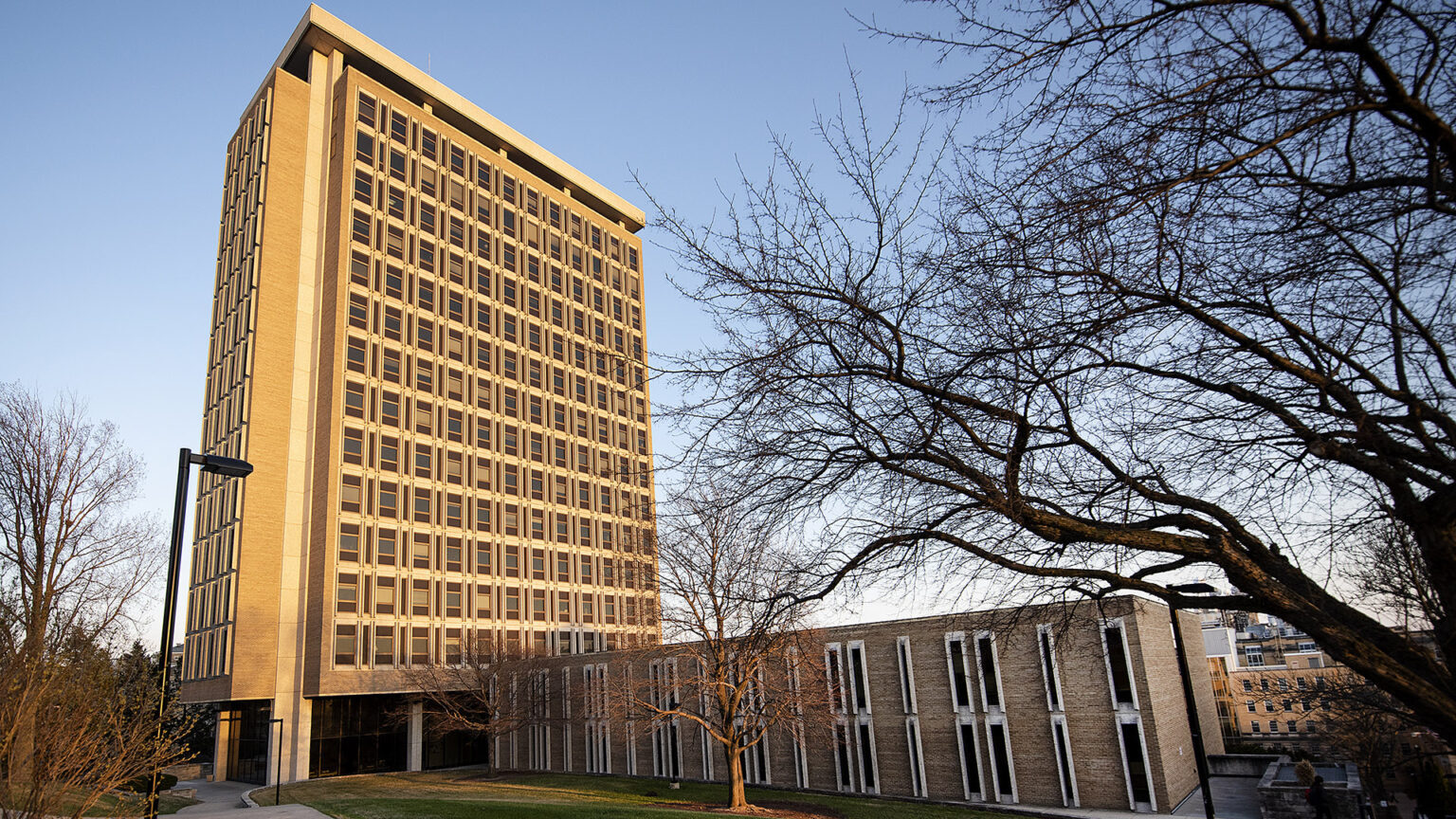 A tall, multi-story portion of a building stands next to a short, two-story wing, with leafless trees in the foreground.