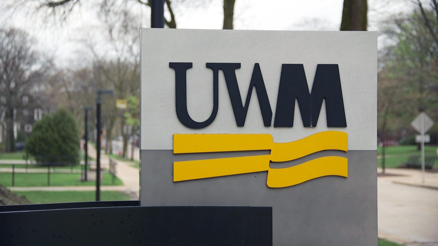 A sign with a UWM wordmark stands next to a road, with trees, buildings and sidewalks in the background.