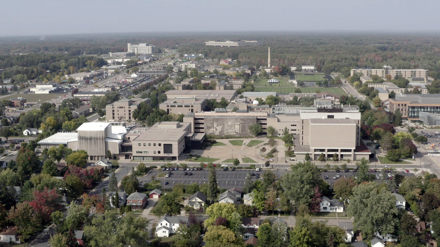 An aerial photo shows a college campus with many trees and variety of structures in the midst of a landscape with residential neighborhoods of houses and wooded areas on the horizon.