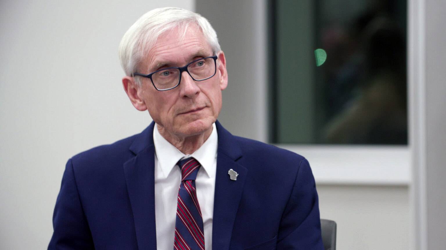 Tony Evers sits in a room with a window in the background and listens to another speaker.