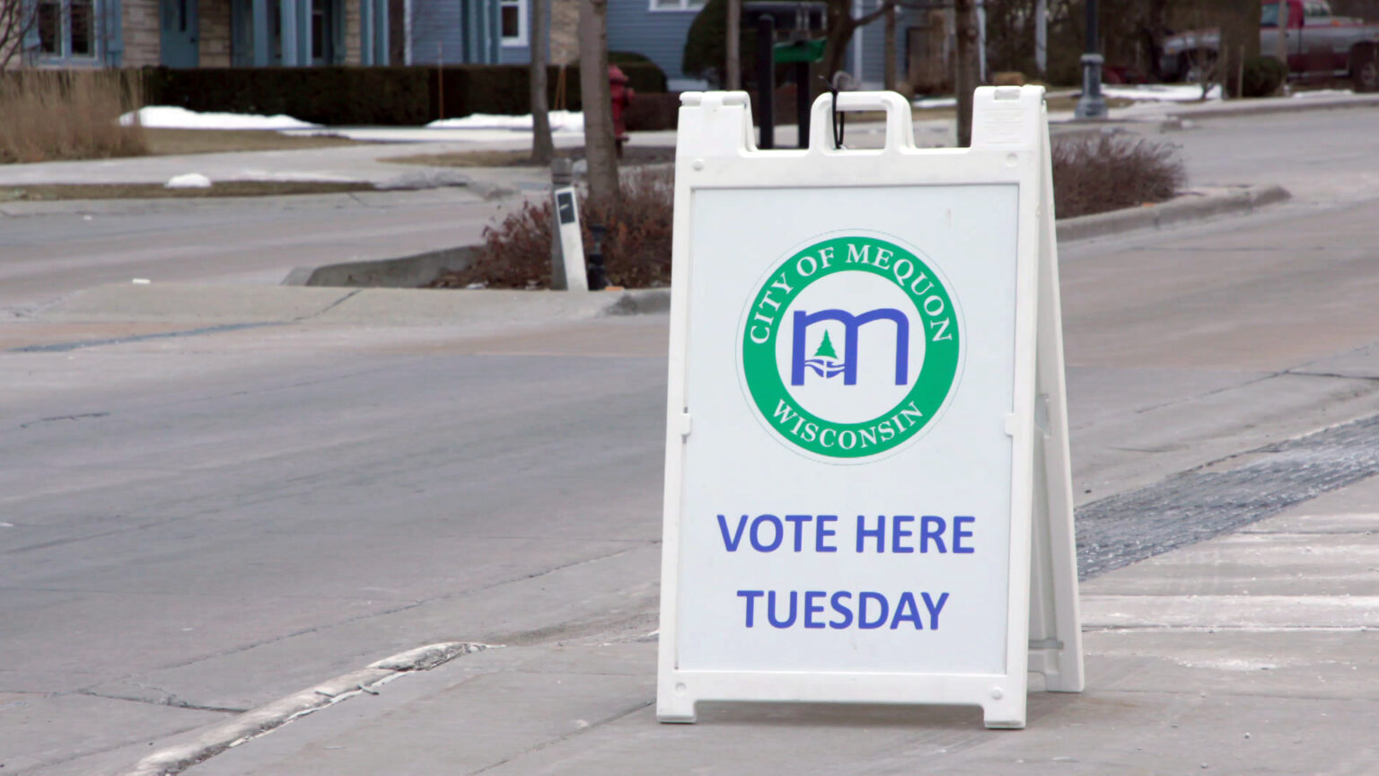 A sandwich board sign with the logo of the city of Mequon and the words Vote Here Tuesday stands on a sidewalk, with a street, trees and buildings in the background.