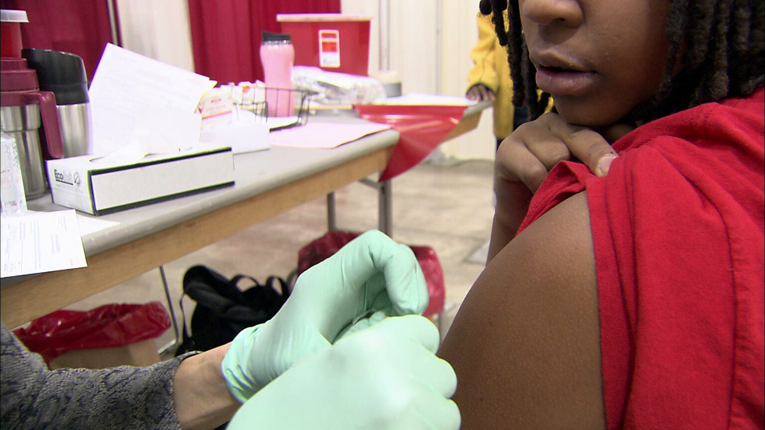 A seated student holds up their left sleeve while another person wearing sterile gloves prepares to administer a vaccine, with a table covered with medical supplies, insulated drinking mugs and other items in the background.