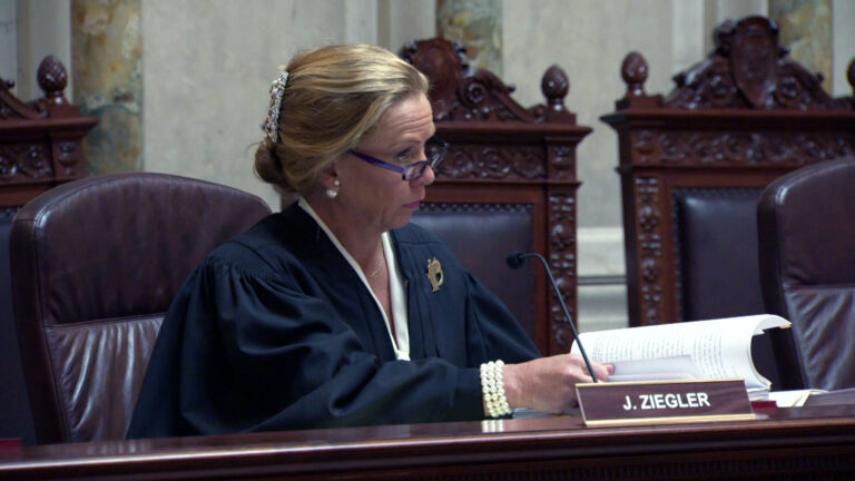 Annette Ziegler sits in a high-backed leather chair at a judicial bench equipped with a microphone and a nameplate reading J. Ziegler, holding a sheaf of papers in her hands while listening to another person speaking, with high-backed carved wood-and-leather chairs in the background and in a room with marble pillars and masonry.