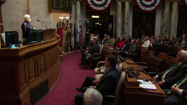 Robert VanZile stands behind a podium on a wood dais and speaks to people seated in rows of rolling chairs and desks, in a meeting room with marble walls and pillars, decorated with eagle staffs and red, white and blue bunting.
