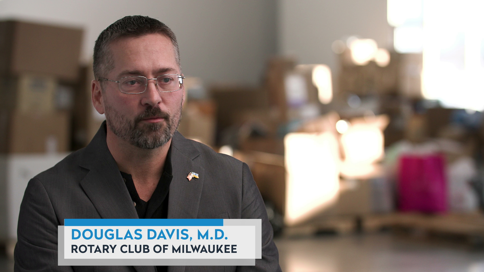 Douglas Davis sits in a large room with out-of-focus stacks of cardboard boxes and other items in the background, with a graphic at bottom reading "Douglas Davis, M.D." and "Rotary Club of Milwaukee."