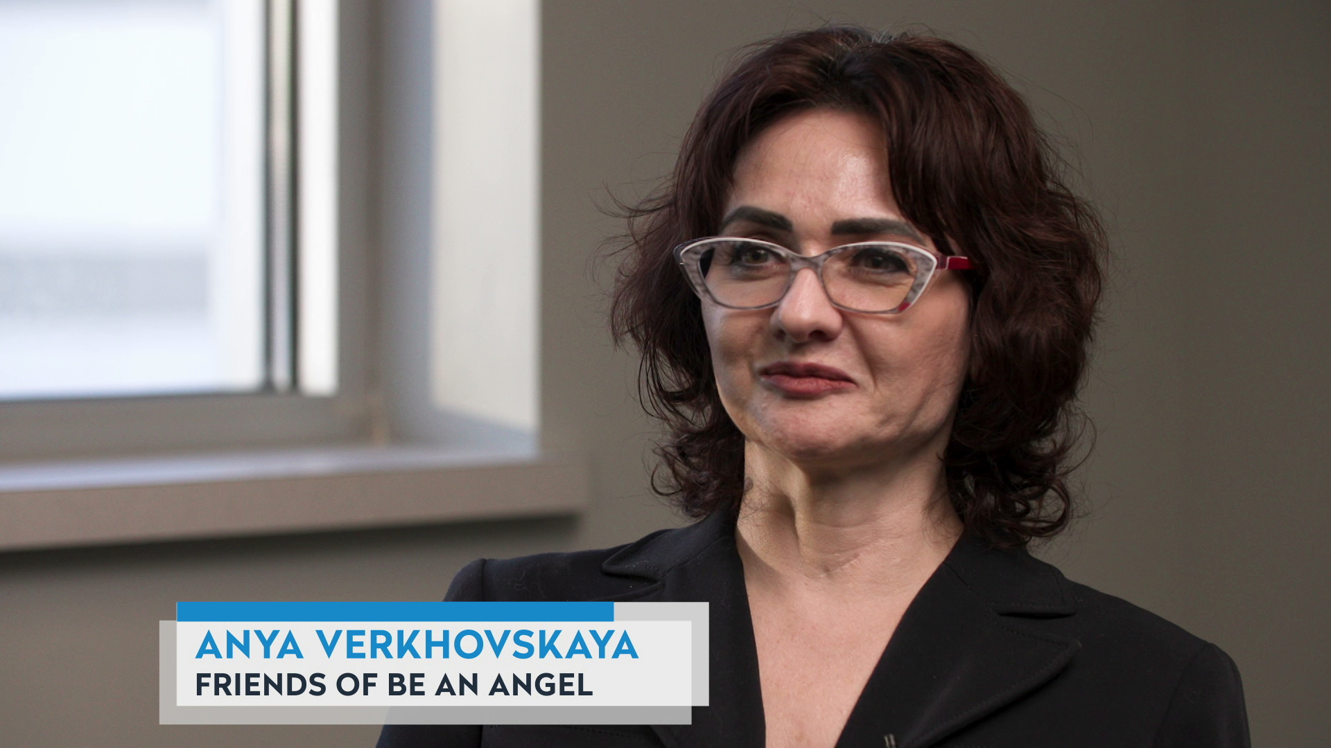Anya Verkhovskaya sits in a room with a window behind her right shoulder, with a graphic at bottom reading "Anya Verkhovskaya" and "Friends of Be an Angel."
