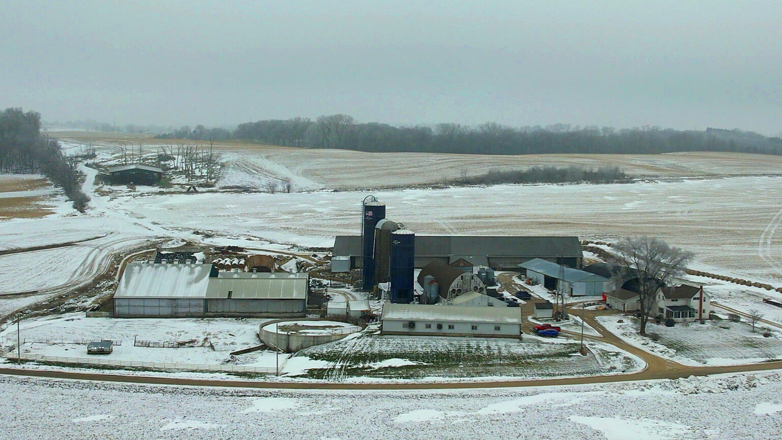 A cluster of farm buildings, including a house, multiple barns and silos, and other outbuildings stand in the midst of fields covered in light snow, with a tree-lined ridge in the background.