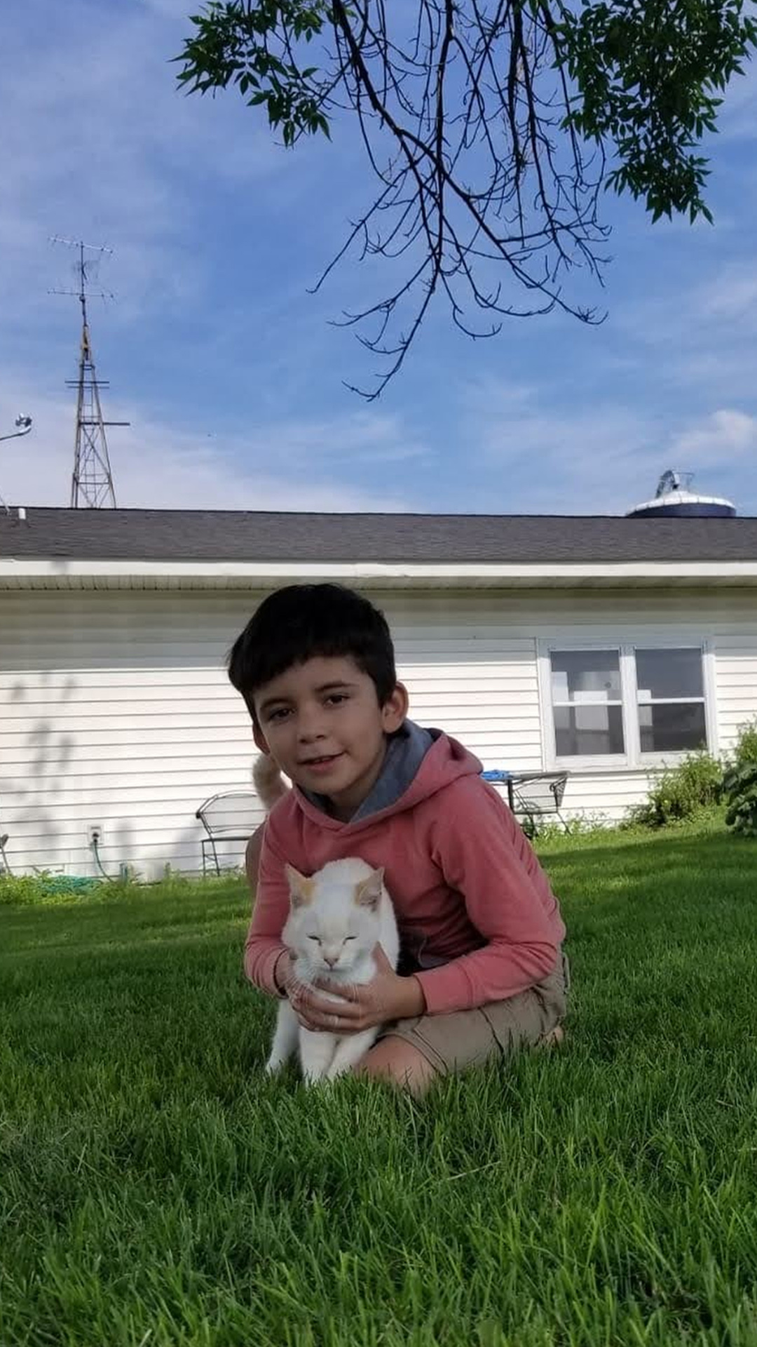 Jefferson Rodríguez sits on a lawn while holding a cat, with a single-story building with a window, radio tower and silo in the background.