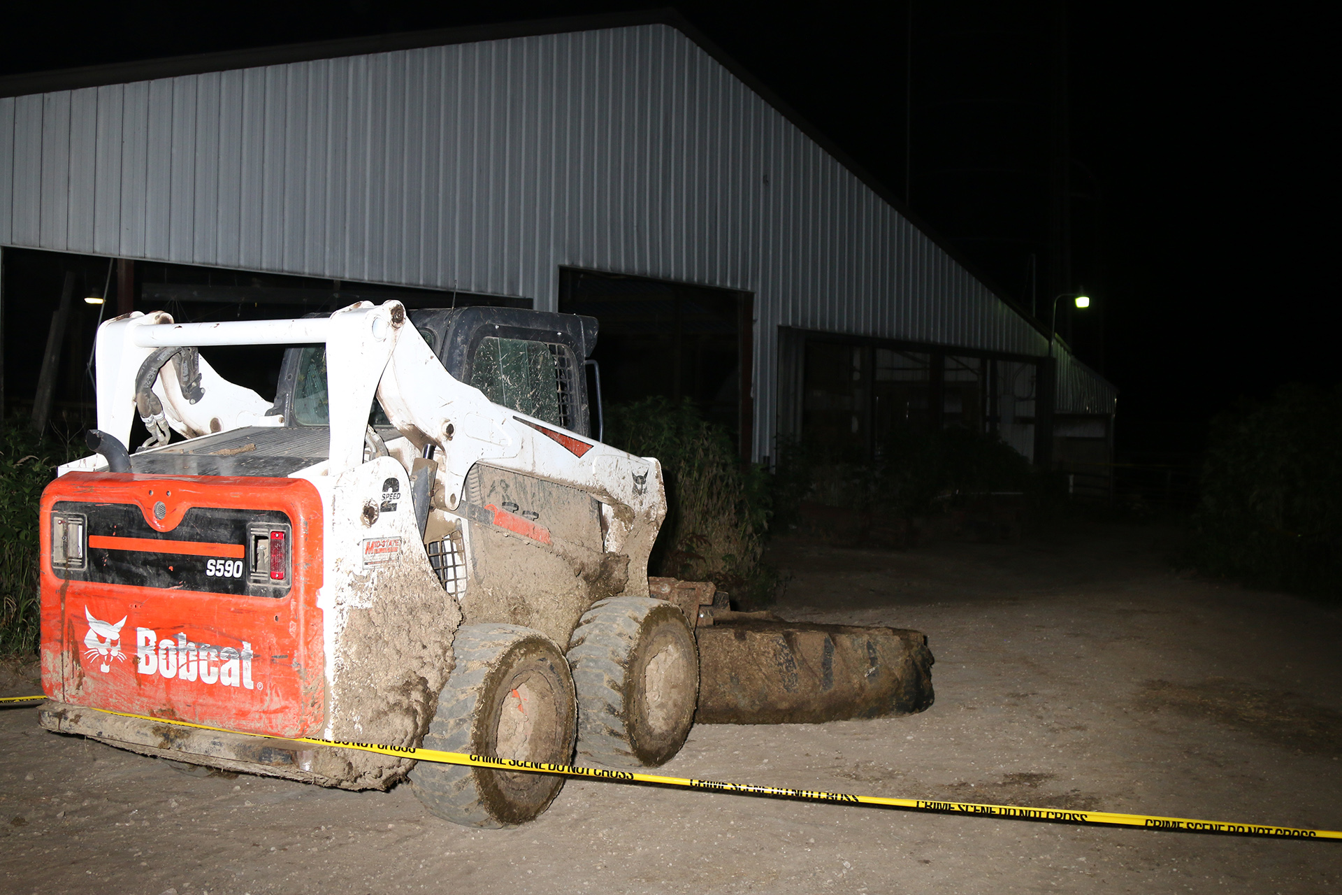 A four-wheeled agricultural vehicle with a front-mounted implement is covered in dirt and manure while parked in front of a metal-sided farm building, with barricade tape reading "Crime Scene" and "Do Not Cross" in the foreground.