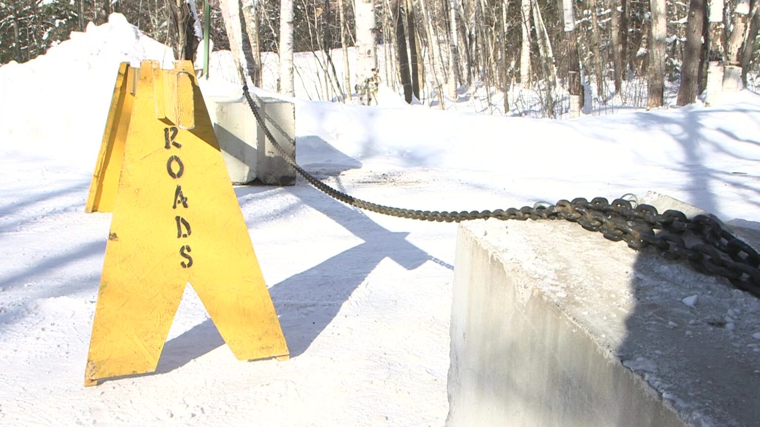 A wood road barrier with Roads painted on its side and two concrete blocks connected with a metal chain sit in front of a road covered in snow, with trees in the background.