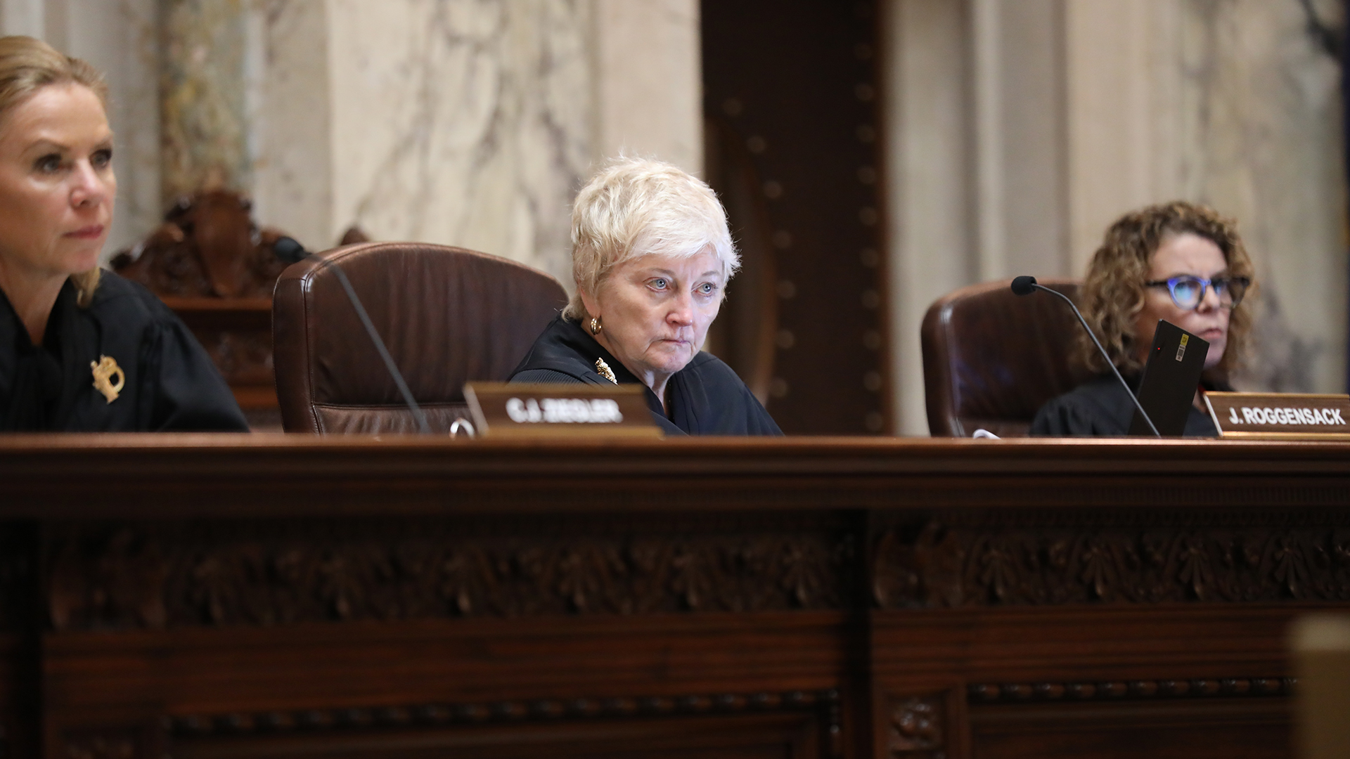 Patience Roggensack sits in a chair behind a judicial bench and listens with two other justices seated on either side of her, in a room with marble walls.