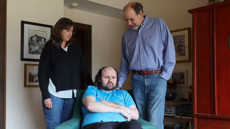 Frank Wallitsch sits in an armchair while Susan and Mark Wallitsch stand behind him, in a room with an armoire, shelves and framed photos on the walls.