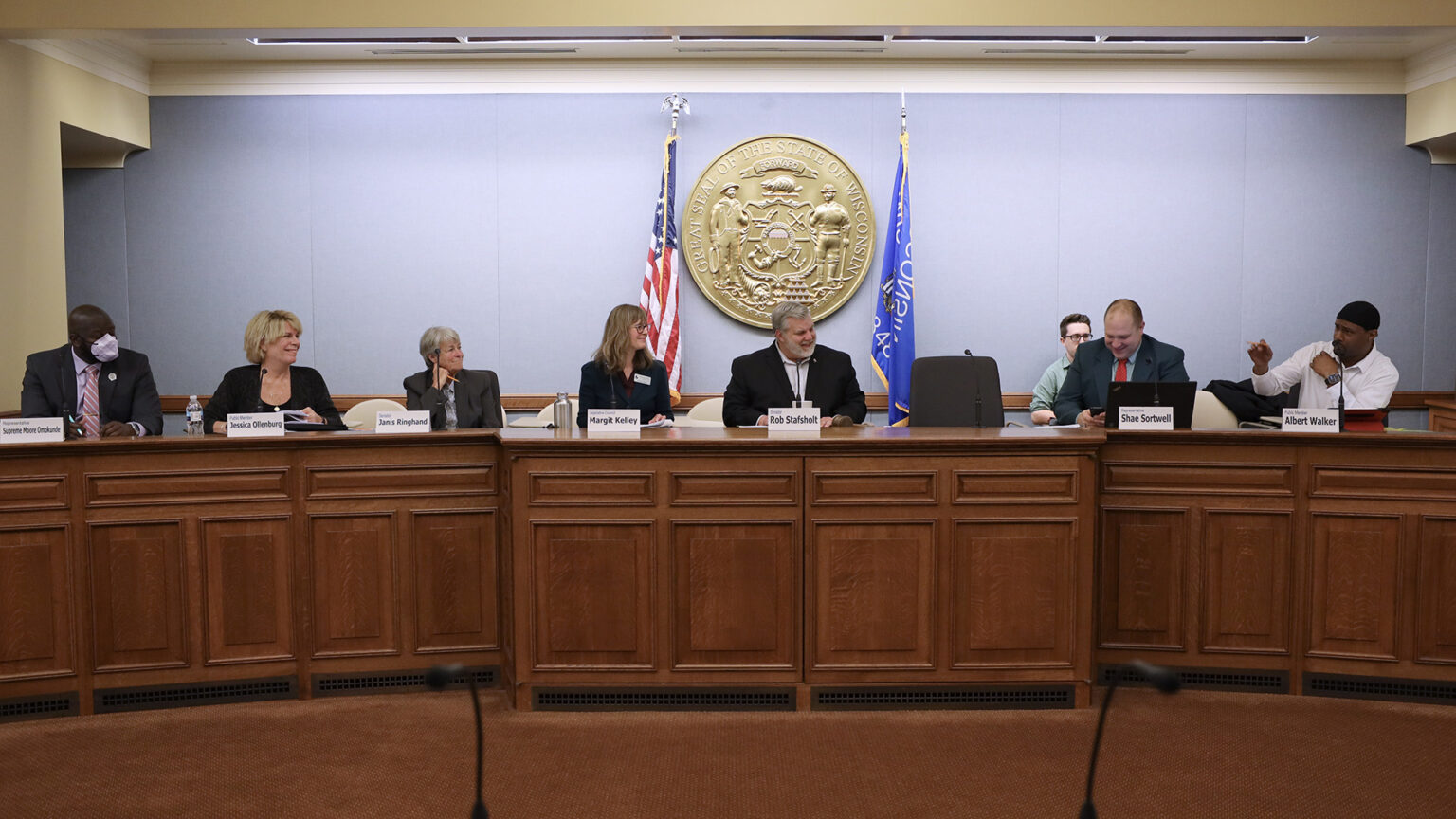 Members of a committee sit in chairs behind a bench desk, with paper name labels and desk-mounted microphones in front of them, and the seal of the state of Wisconsin mounted on the wall in the background with the U.S. and Wisconsin flags standing on either side.