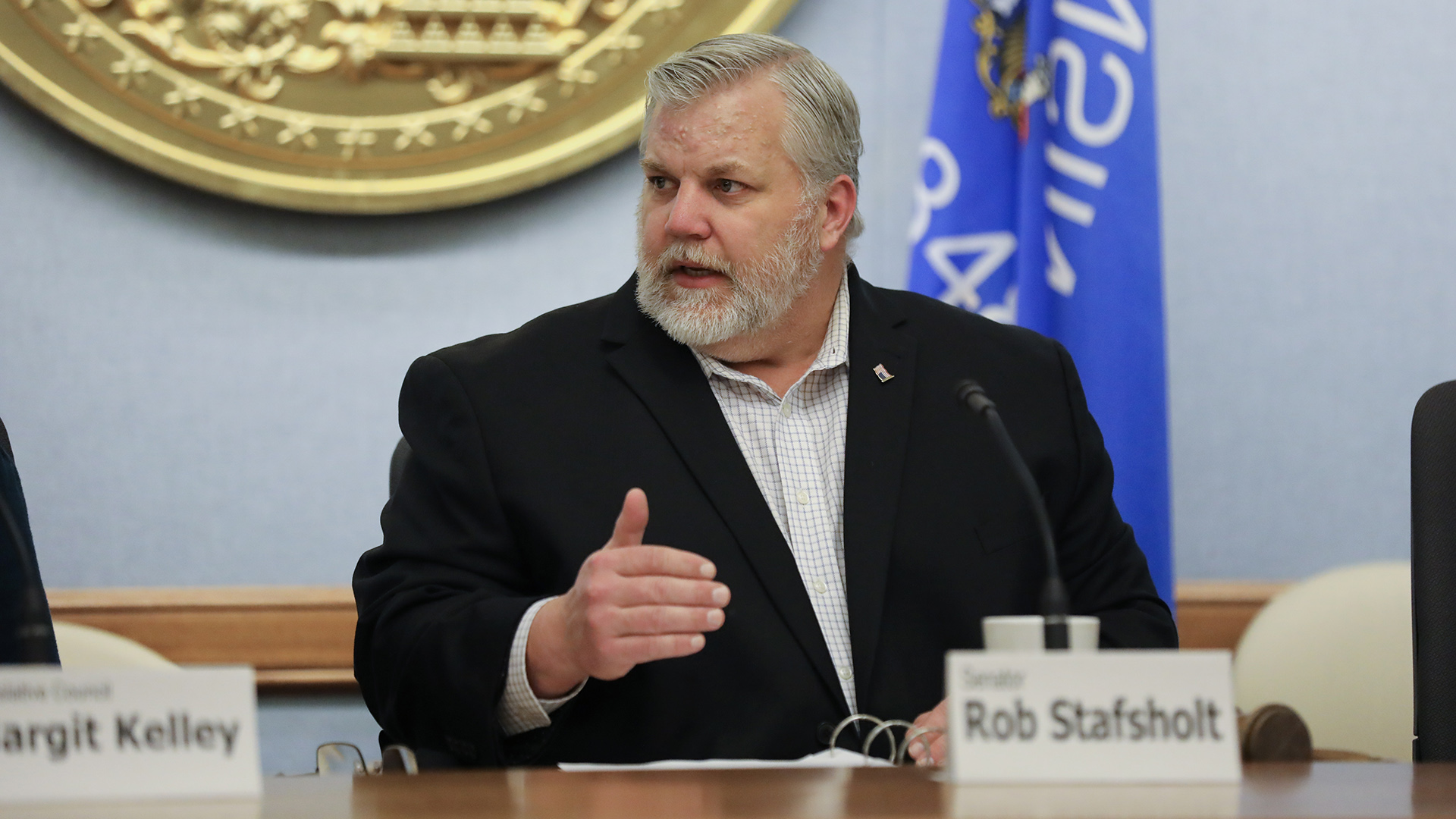 Rob Stafholt sits at a desk and speaks while gesturing with his right hand, with a paper name label and mounted microphone in front of him and a Wisconsin flag in the background.
