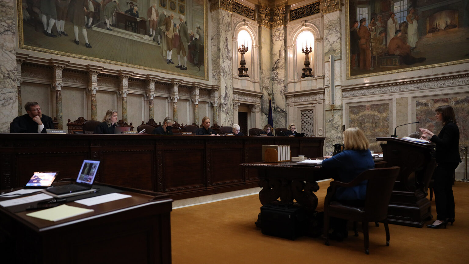 A person standing behind a wood podium, with another person seated to her left at a wood table, speaks in a microphone toward a wood judicial dais with seven seated justices, in a room with marble pillars and masonry, electric sconces and large paintings.