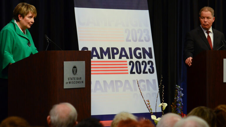 Janet Protasiewicz stands behind a podicum with a metal sign showing the wordmark for the State Bar of Wisconsin and speaks into a microphone while Daniel Kelly stands behind another podium and listens, with a stage curtain and sign reading Campaign 2023 in the background and the out-of-focus heads of an audience in the foreground.