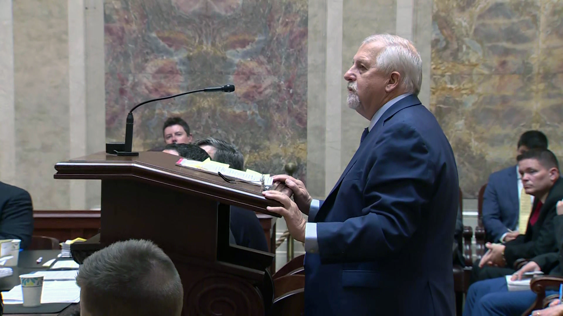 Rick Esenberg stands behind a wood podium with a microphone and speaks with other people seated in front of and behind him, in a room with marble masonry.