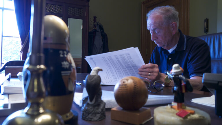 N. Patrick Crooks sits at a desk and pages through papers, with a bald eagle statue, commemorative baseball, lamp and other items sitting in the foreground, and an armoire with mirrored doors and window with open drapes in the background.