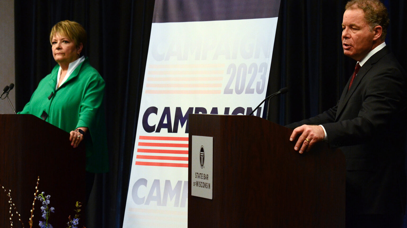 Daniel Kelly stands behind a podicum with a metal sign showing the wordmark for the State Bar of Wisconsin and speaks into a microphone while Janet Protasiewicz stands behind another podium and listens, with a stage curtain and sign reading Campaign 2023 in the background.