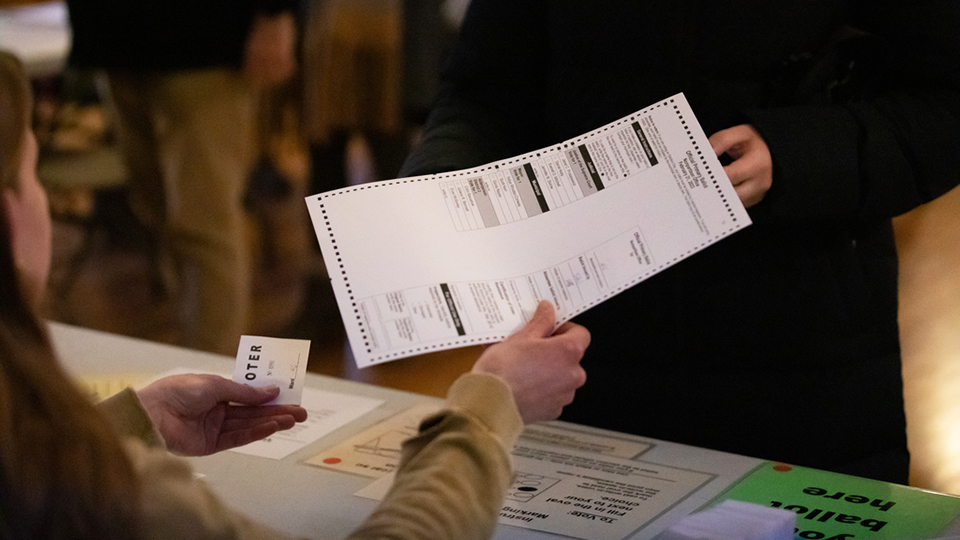A person sitting at a table hands a ballot to another person who is standing and facing them.