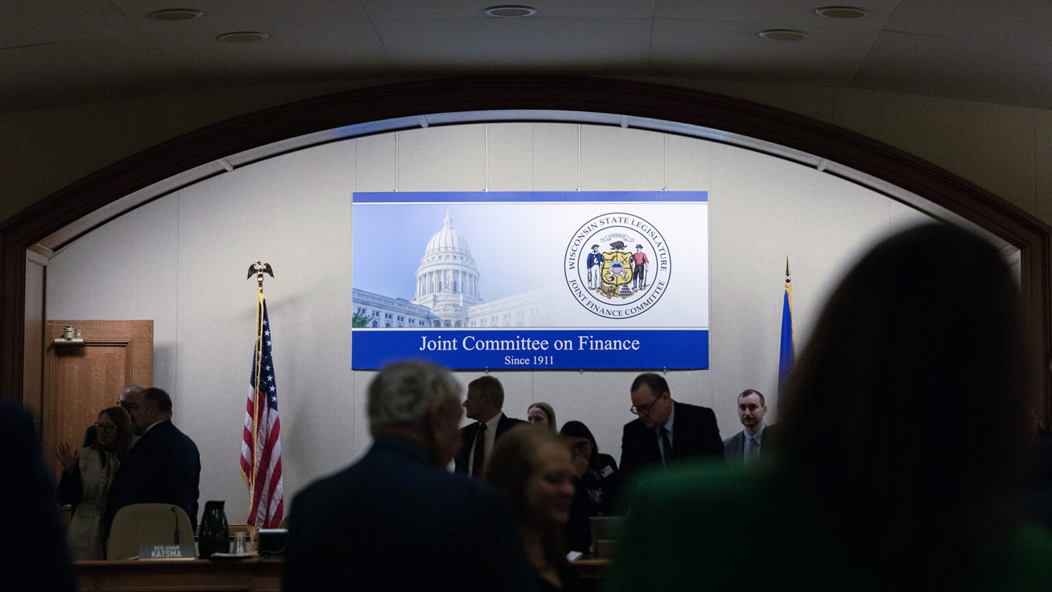 A sign showing a photo of the Wisconsin State Capitol dome and the state seal along with the words Joint Committee on Finance and Since 1911 is mounted on a wall in a room with an arched entryway, with people in shadow standing among desks and the U.S. and Wisconsin flags.