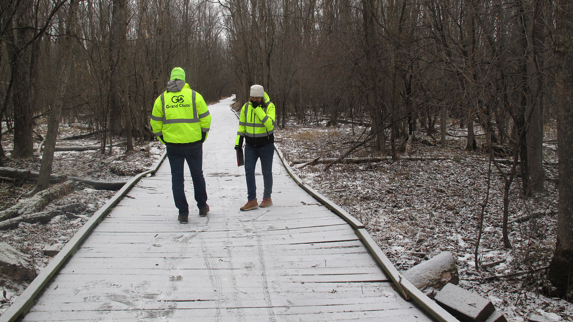 Two people wearing neon reflective jackets stand on a snow-dusted boardwalk passing through a wooded area.