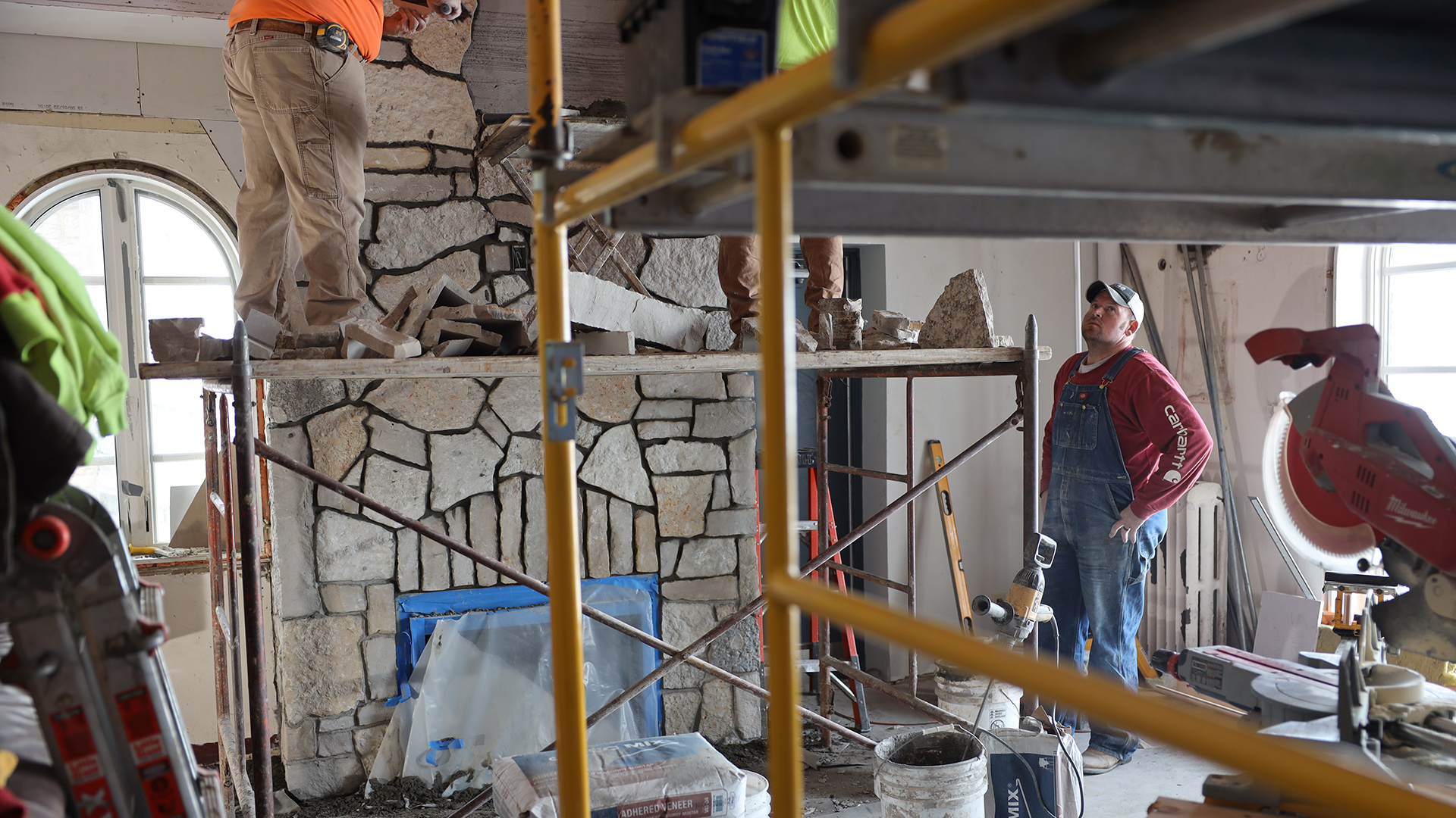 A construction worker stands atop scaffolding in front of a masonry fireplace while another worker watches while standing on the floor, with more scaffolding and other construction equipment and supplies in the room.