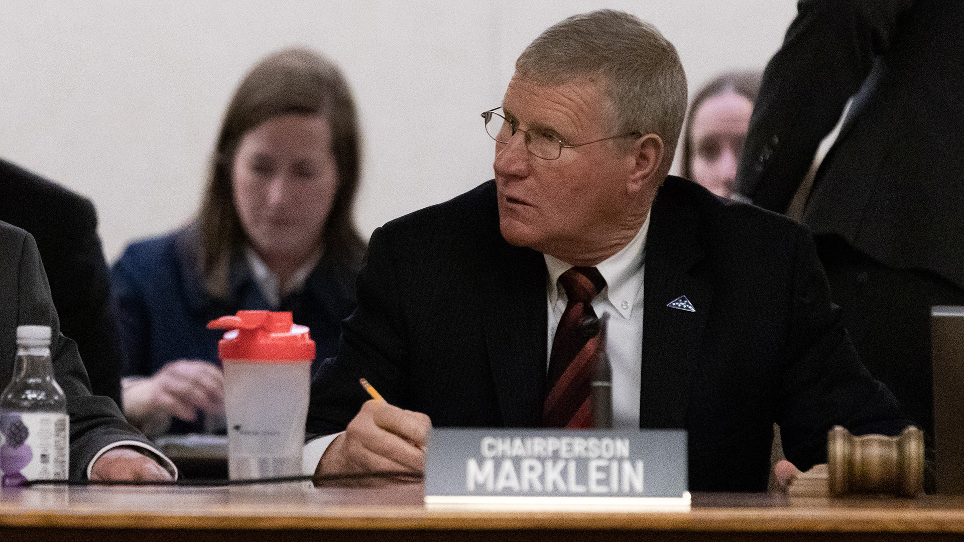 Howard Marklein sits at a table and writes with a pencil in his right hand, with a gavel sitting next to his left hand and a nameplate reading "Chairperson Marklein," with other people seated and standing in the background.