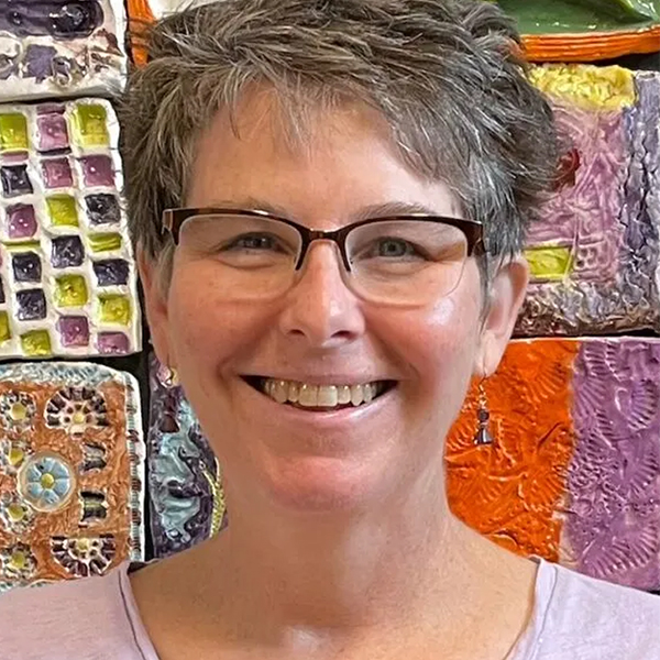 Woman (Virginia Harrison) wearing glasses and standing in front of colorful patchwork.