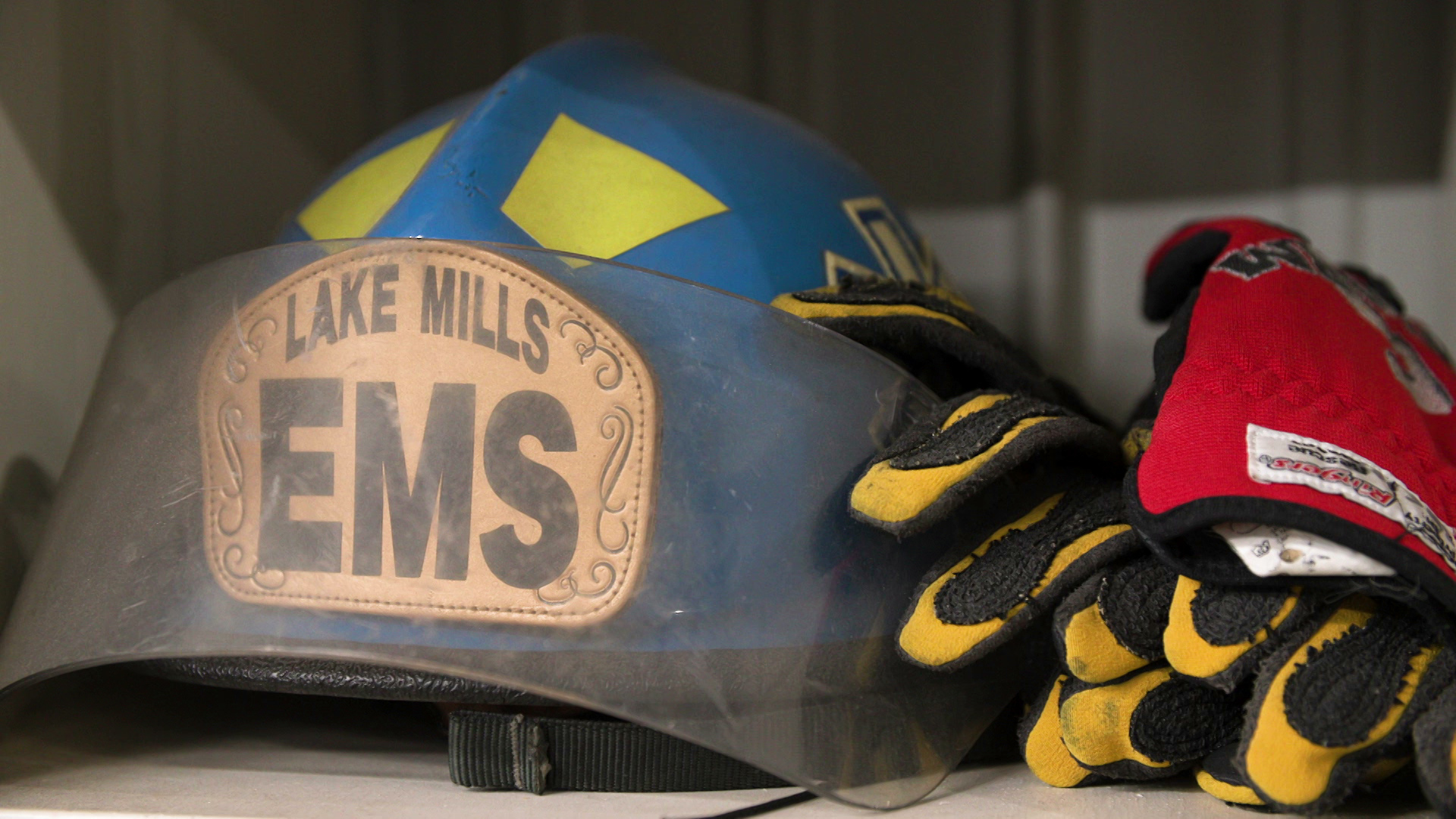 A fire helmet with "Lake Mills EMS" printed in large text on its front sits on a shelf alongside with a pair of padded safety gloves