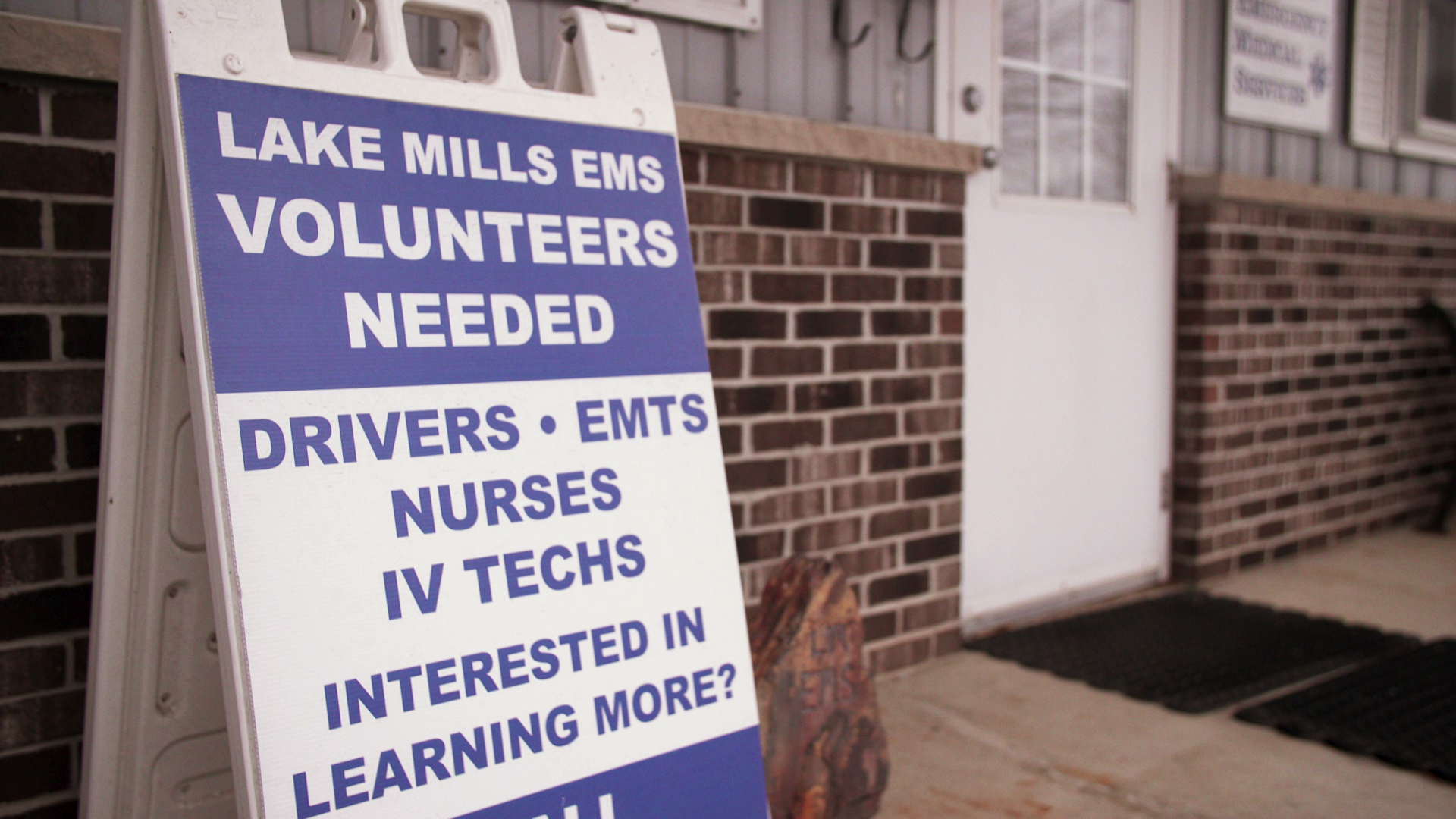 A sandwich board sign placed near a door of a building with brick and wood siding reads "Lake Mills EMS Volunteers Needed," "Drivers • EMTs • Nurses • IV Techs • Interested in Learning More?"