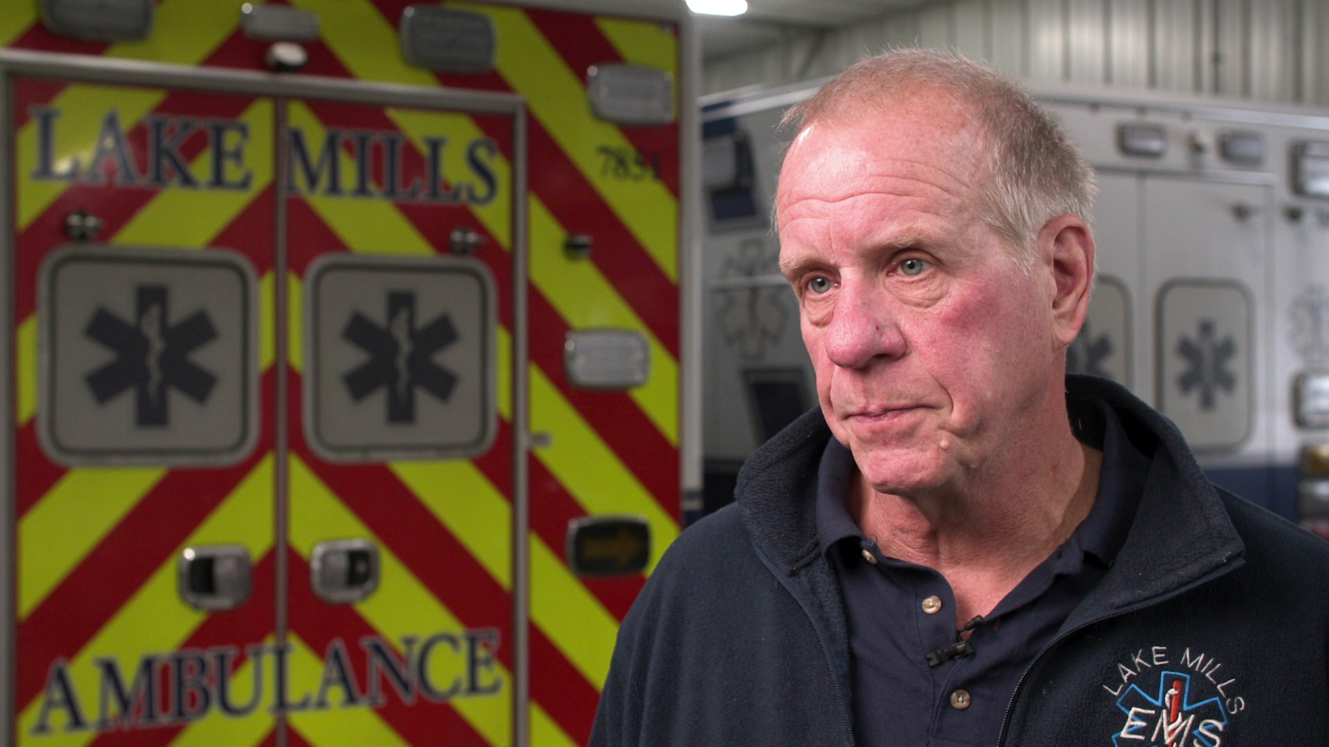 A still image from a video shows Jim Colegrove sitting in a garage with two ambulances in the background.