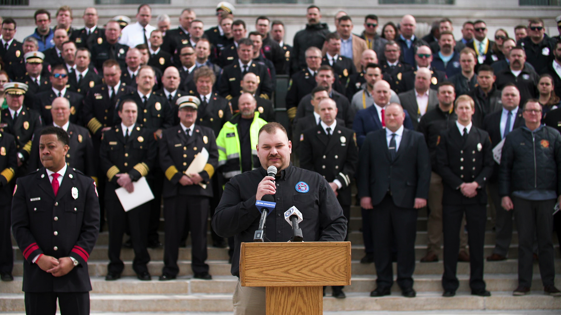 Alan DeYoung speaks into a microphone while standing behind a podium on the Wisconsin State Capitol steps, with dozens EMS workers and other first responders standing behind him.