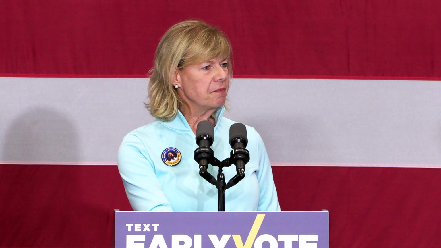 Tammy Baldwin speaks into microphones behind a podium, with the stripes of a large U.S. flag in the background.