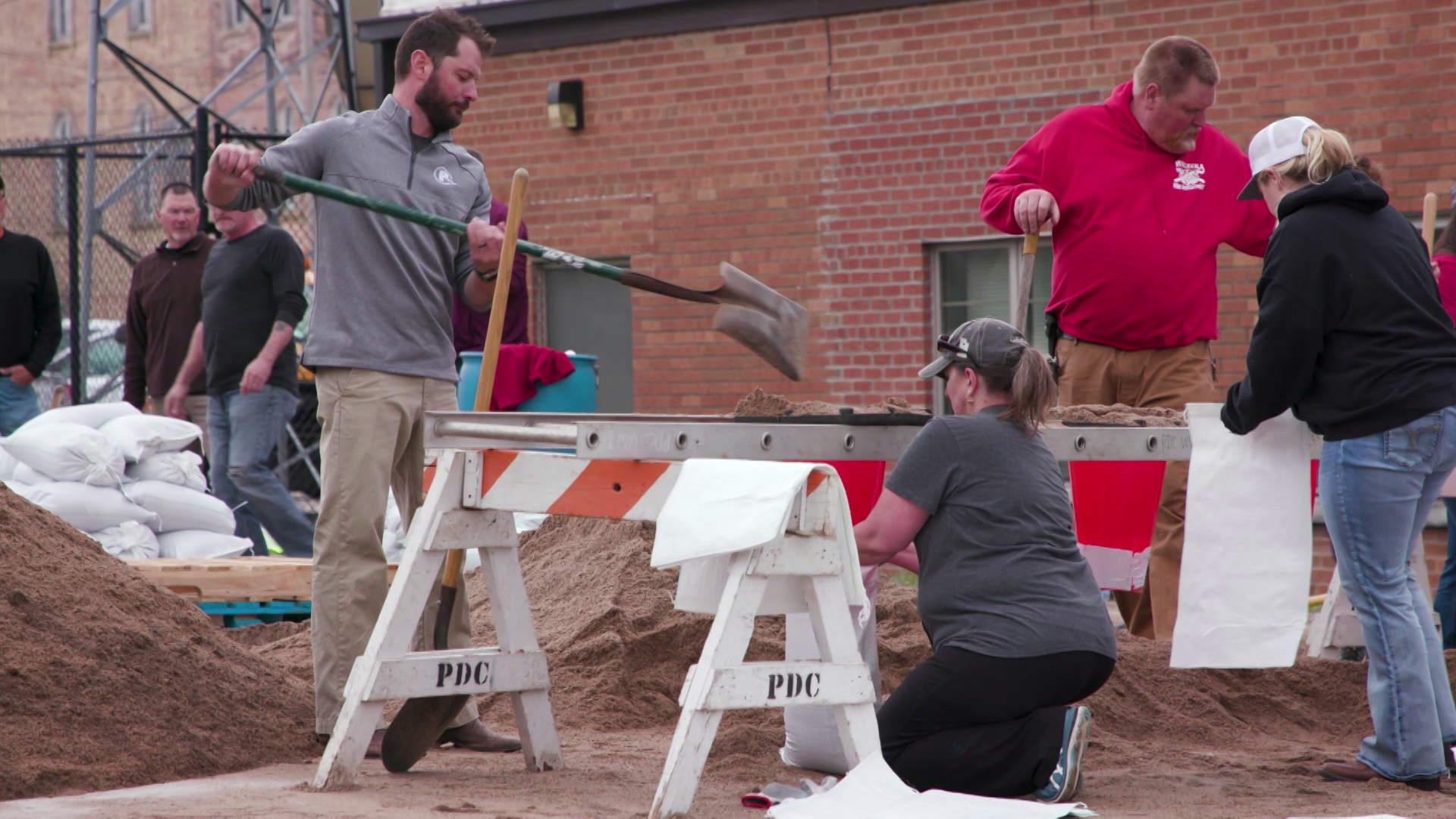 Multiple people stand and crouch among piles of sand as they use shovels to fill sandbags next to a brick building.