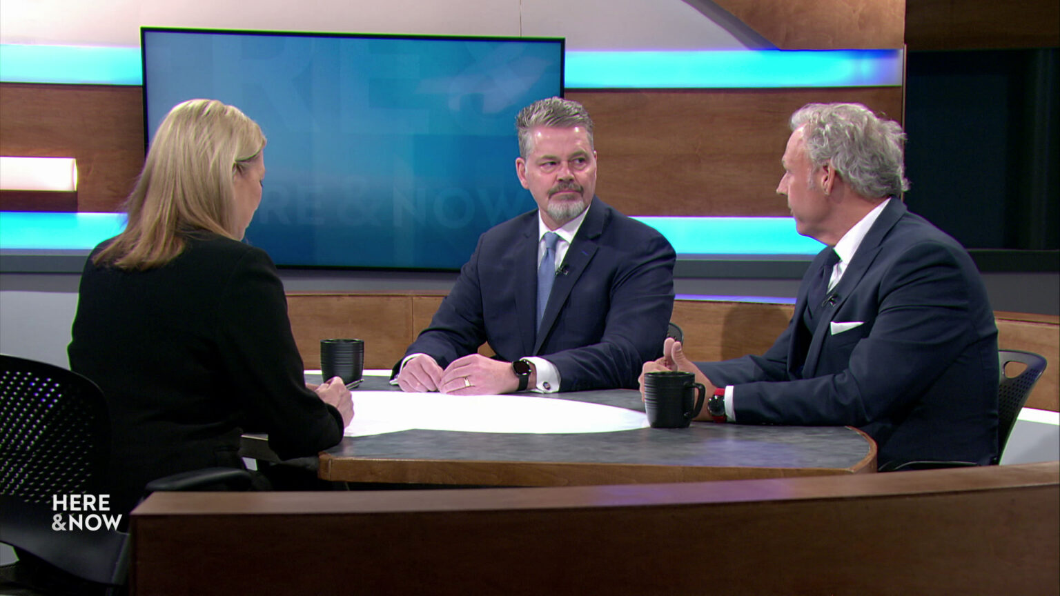 Bill McCoshen and Scot Ross sit next to each other and across from Frederica Freyberg at a desk on the Here & Now set.