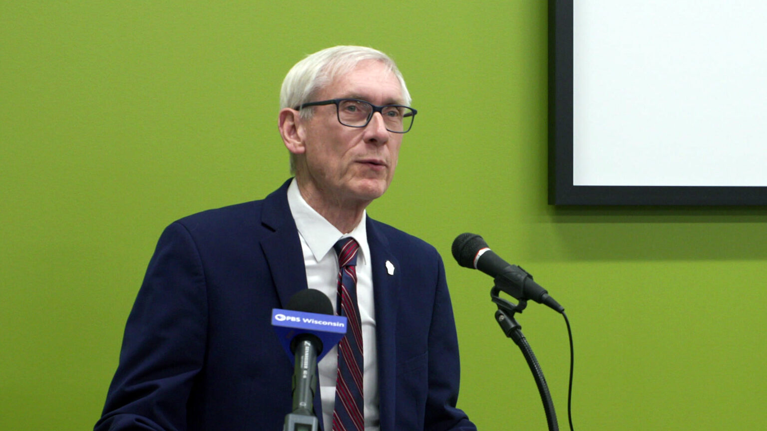 Tony Evers stands and speaks into two microphones, with a whiteboard on the wall behind him.