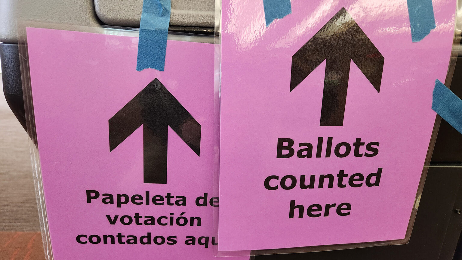 Two laminated signs with arrows pointing up read Papeleta de votación contados aqui and Ballots counted here are attached with painter's tape to the side of a ballot tabulator machine.