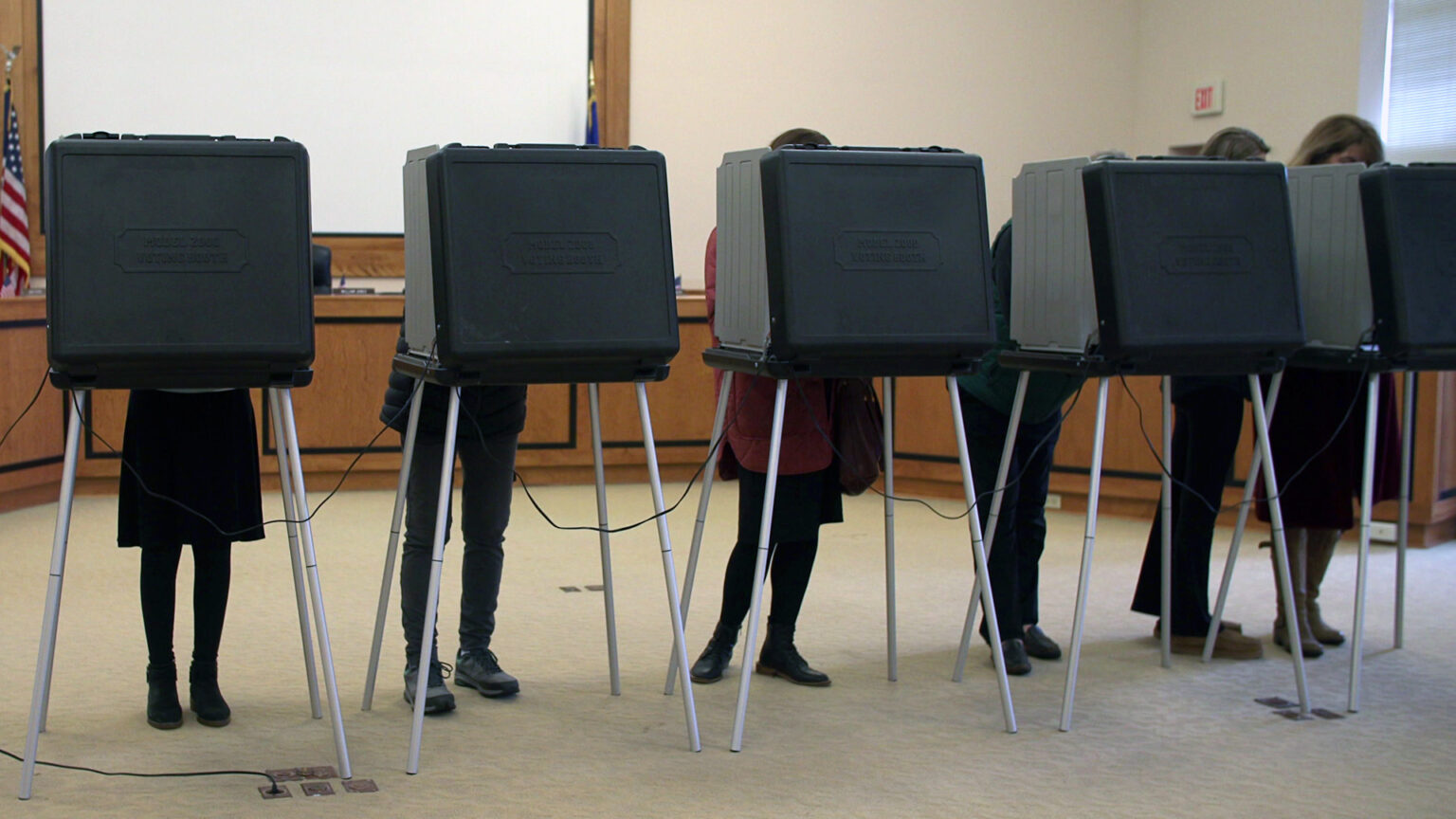 Six voters stand and face the interior of temporary voting privacy booths with plastic frames and metal legs, in a government meeting room with a row of desks, projector screen and U.S. flag in the background.