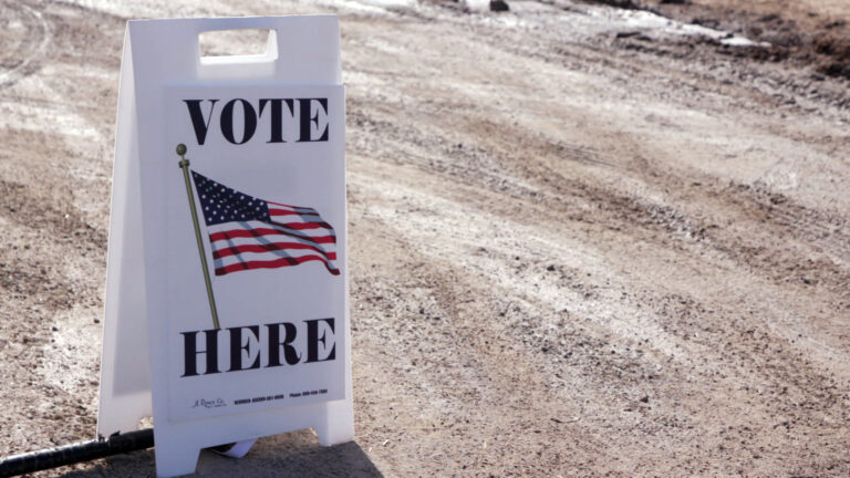 A poster with a U.S. flag graphic and the words Vote Here is affixed to a sandwich board sign standing on a gravel driveway.